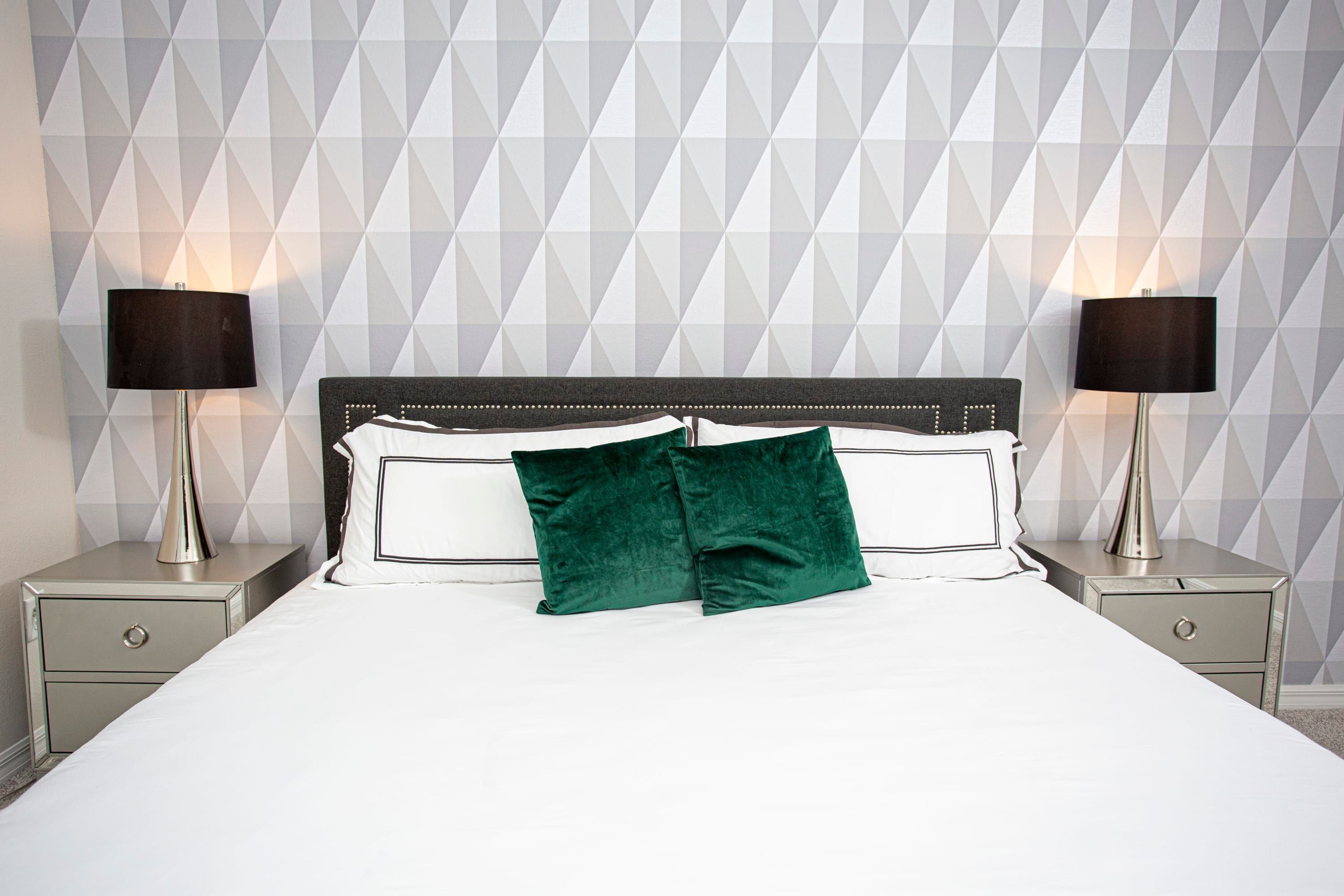 A neatly made bed with a white comforter and two pillows, flanked by two bedside tables with lamps, against a geometric-patterned wall.