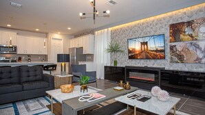 A modern living room with a sofa, coffee table, and fireplace. There is a kitchen in the background with white cabinets and appliances. - Fabulous 9BD 6BA Starwarsgamer Sleep22 Solarareso (Kissimmee)