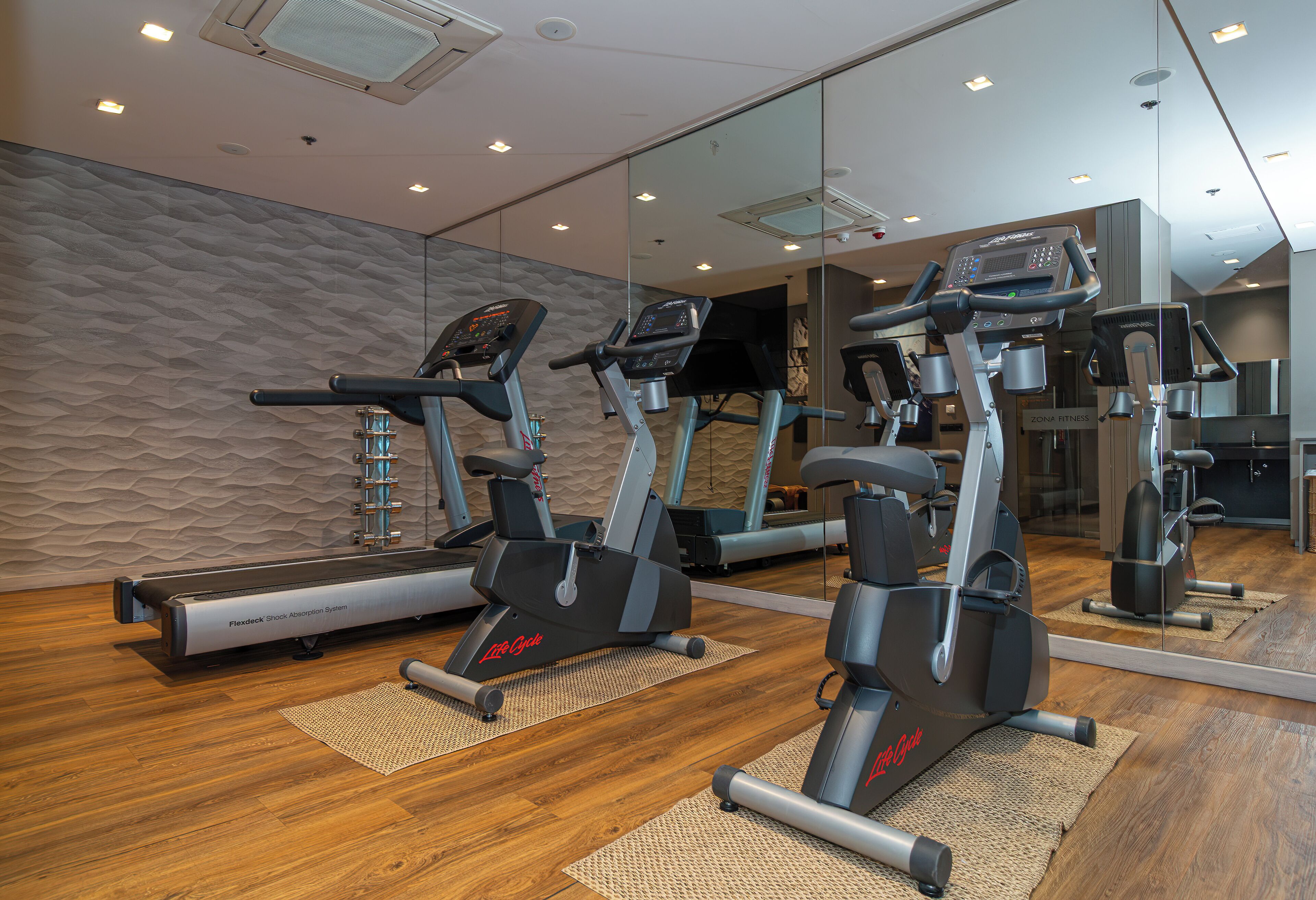 Un gimnasio con tres máquinas de cardio, un espejo fijado a la pared y suelo de madera.