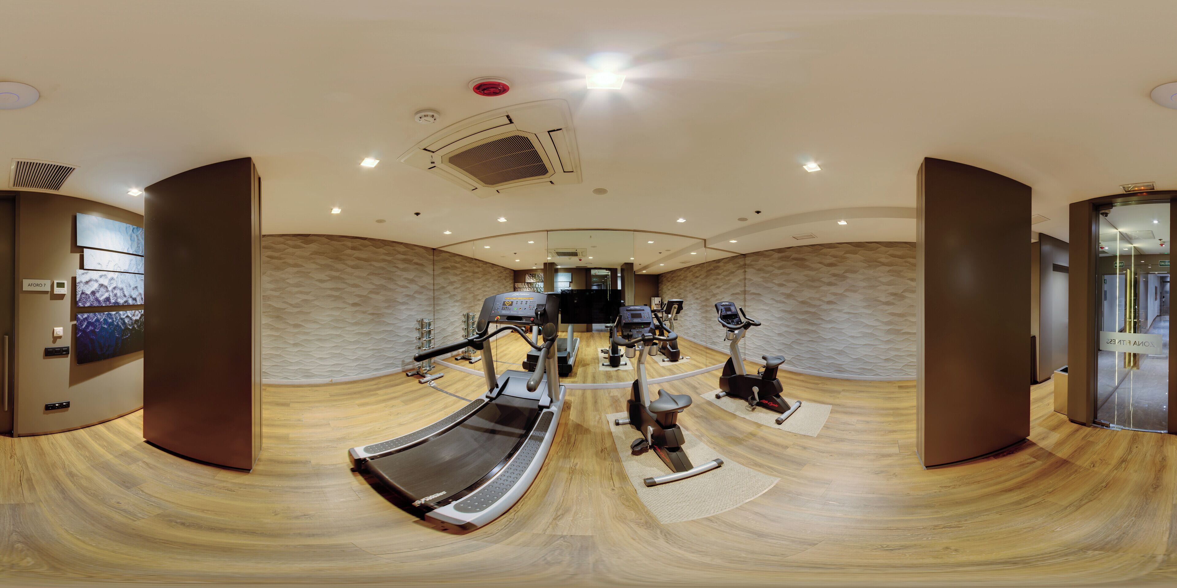 Vista panorámica de un gimnasio con equipo de ejercicio, un espejo grande y suelo de madera.