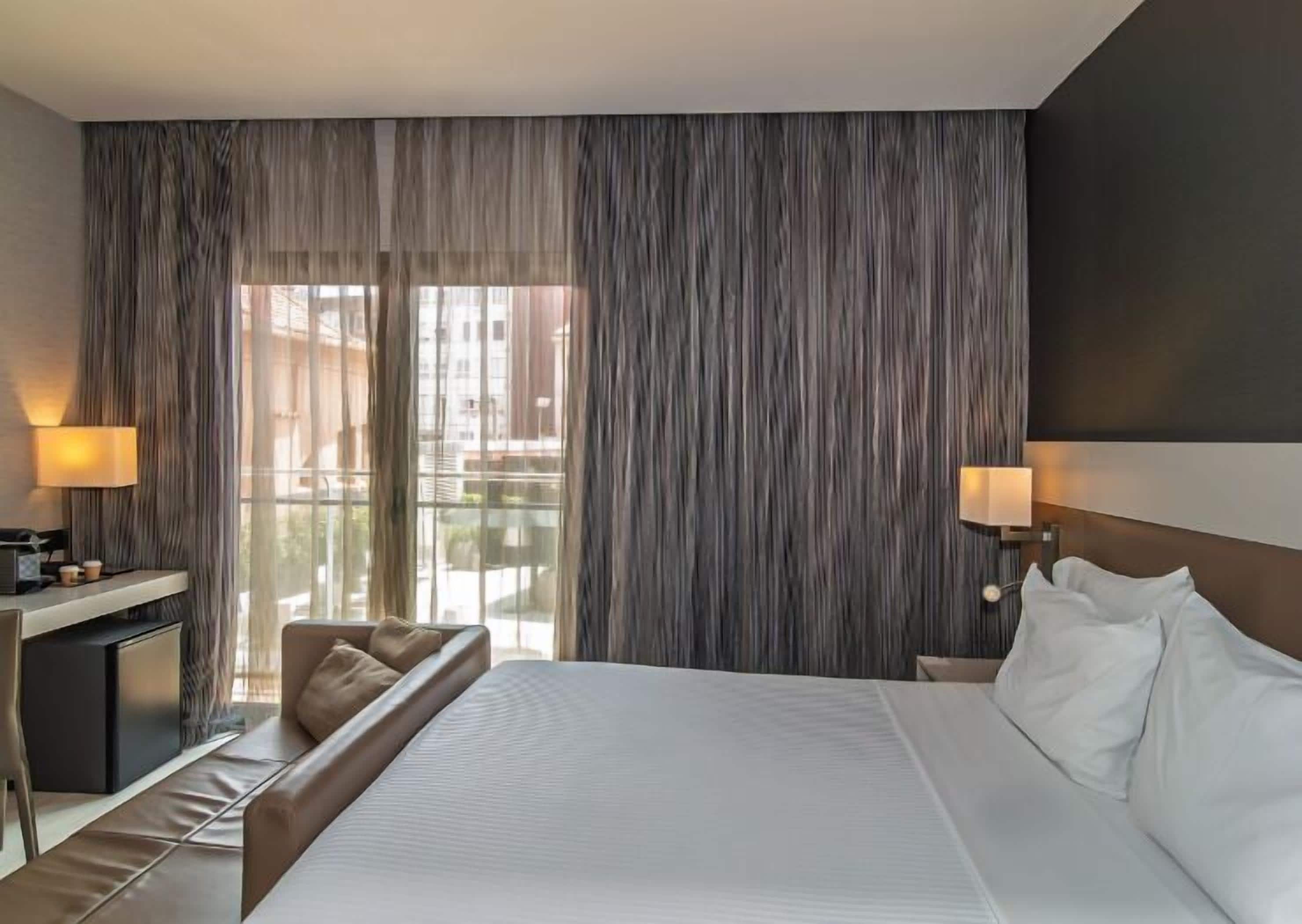 Habitación de hotel con una cama grande, un escritorio con lámpara, una silla y vistas al exterior a través de cortinas traslúcidas.