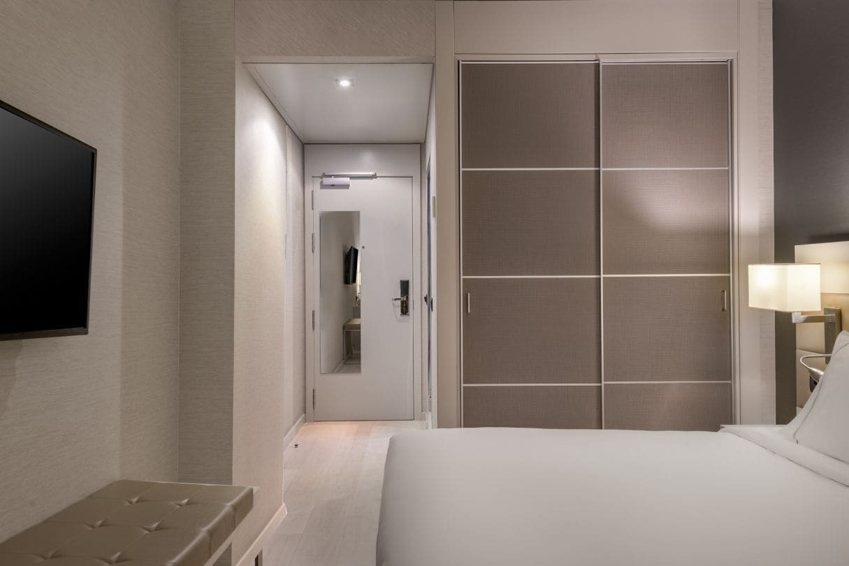 Habitación de hotel con cama, televisor montado en la pared, una banca, armario y puerta que da al baño.
