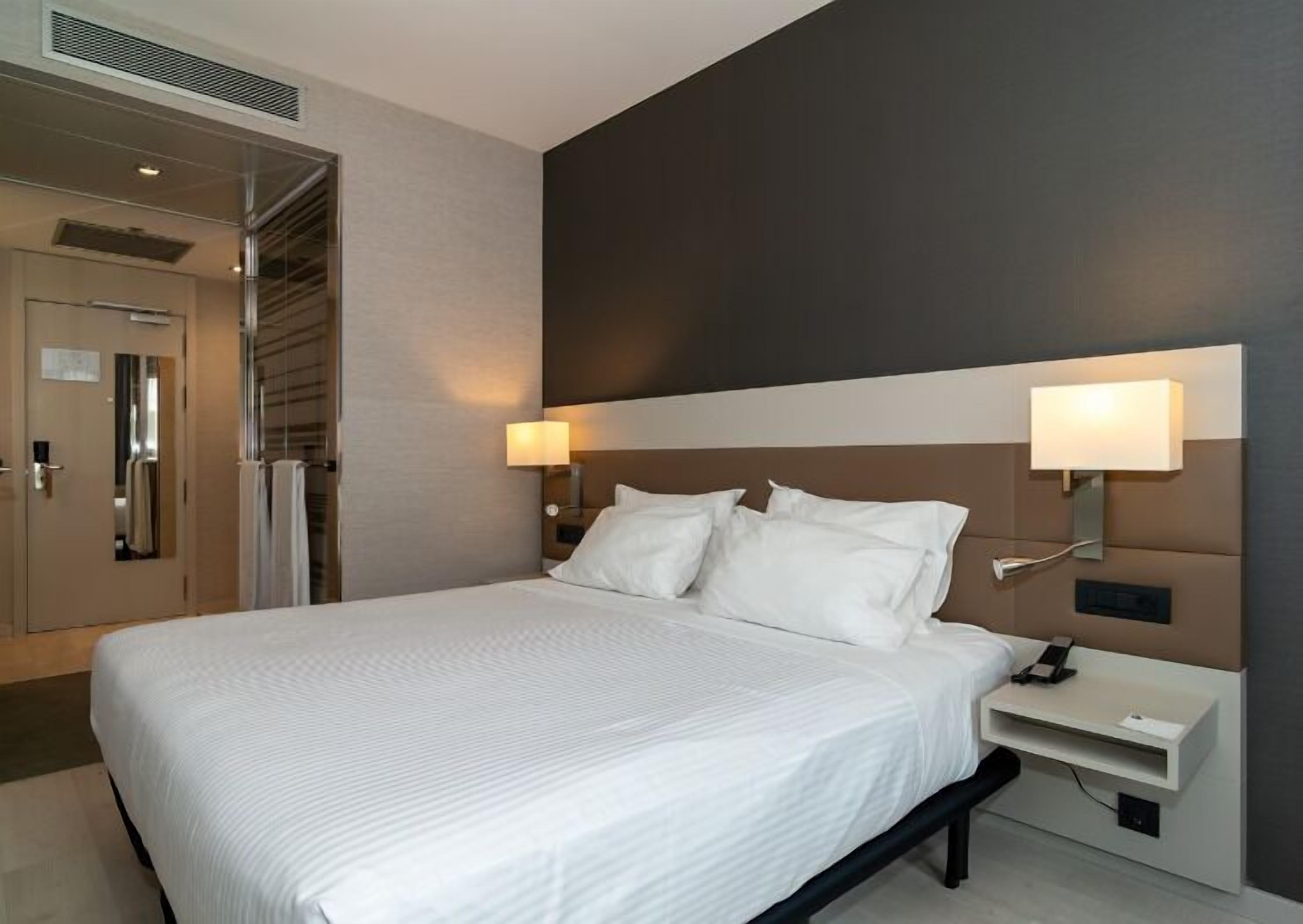 Quarto de hotel com uma cama grande, mesas de cabeceira, uma luminária na parede e uma porta com espelho.