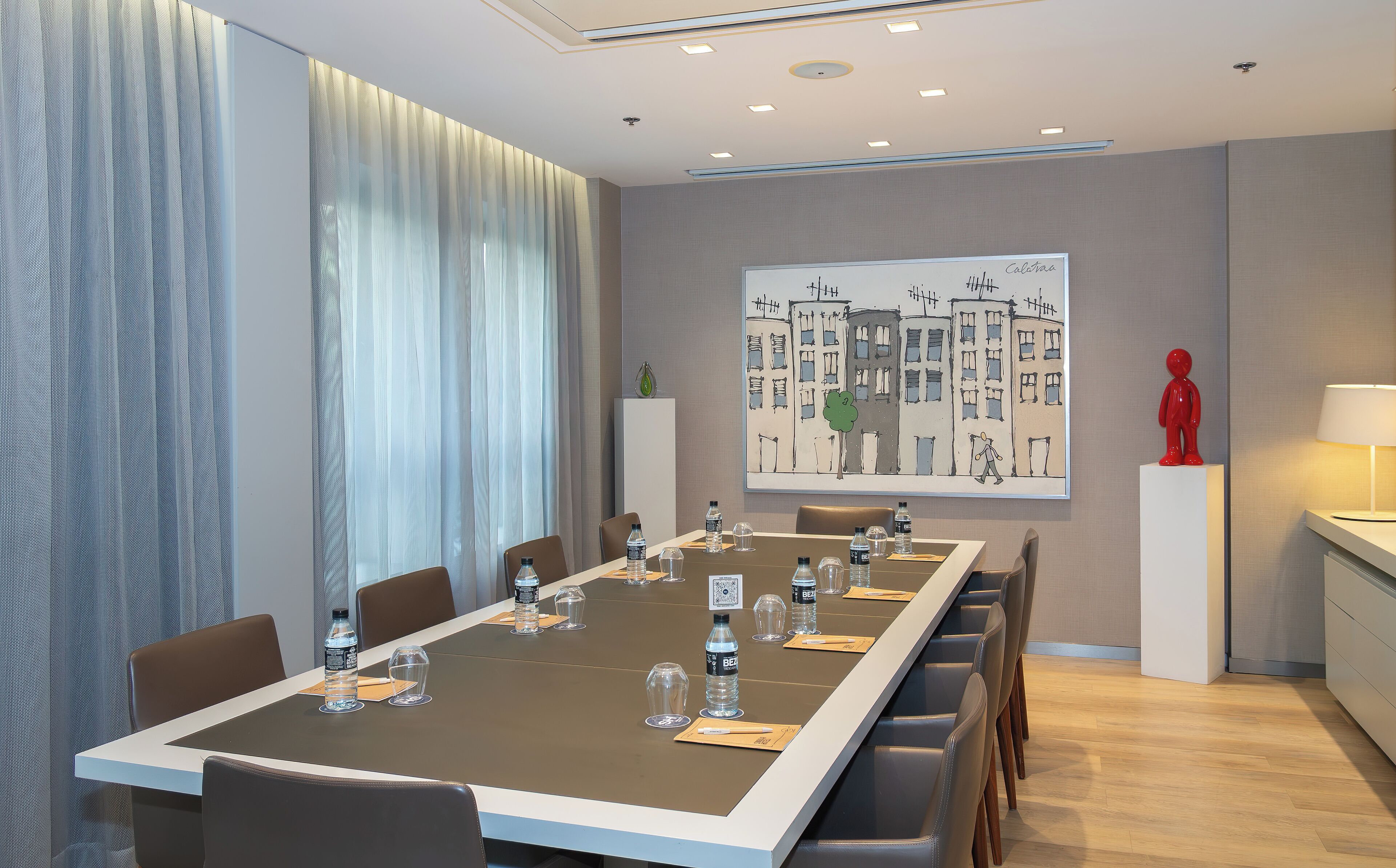 A modern conference room with a long table, chairs, water bottles, and a painting of buildings.