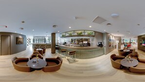 A modern hotel lobby with a curved reception desk, seating areas with round tables, and a large TV screen.