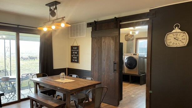 Un coin repas avec une table et des chaises en bois, un luminaire suspendu, une horloge au mur et une vue sur l’extérieur.