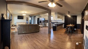 Un vaste espace de vie comprenant un canapé, un plancher en bois, un ventilateur de plafond et une cuisine à l’arrière-plan.