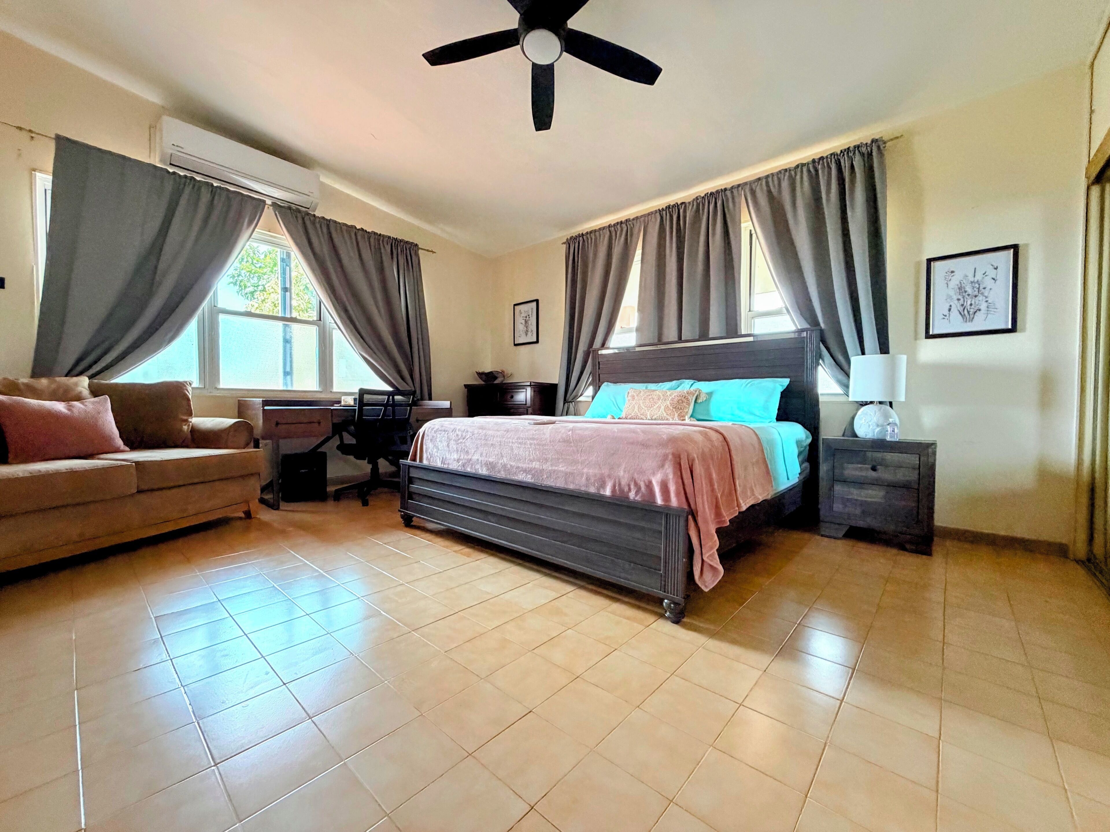 Un dormitorio con cama, escritorio, silla, sofá, ventilador de techo y ventana con cortinas.
