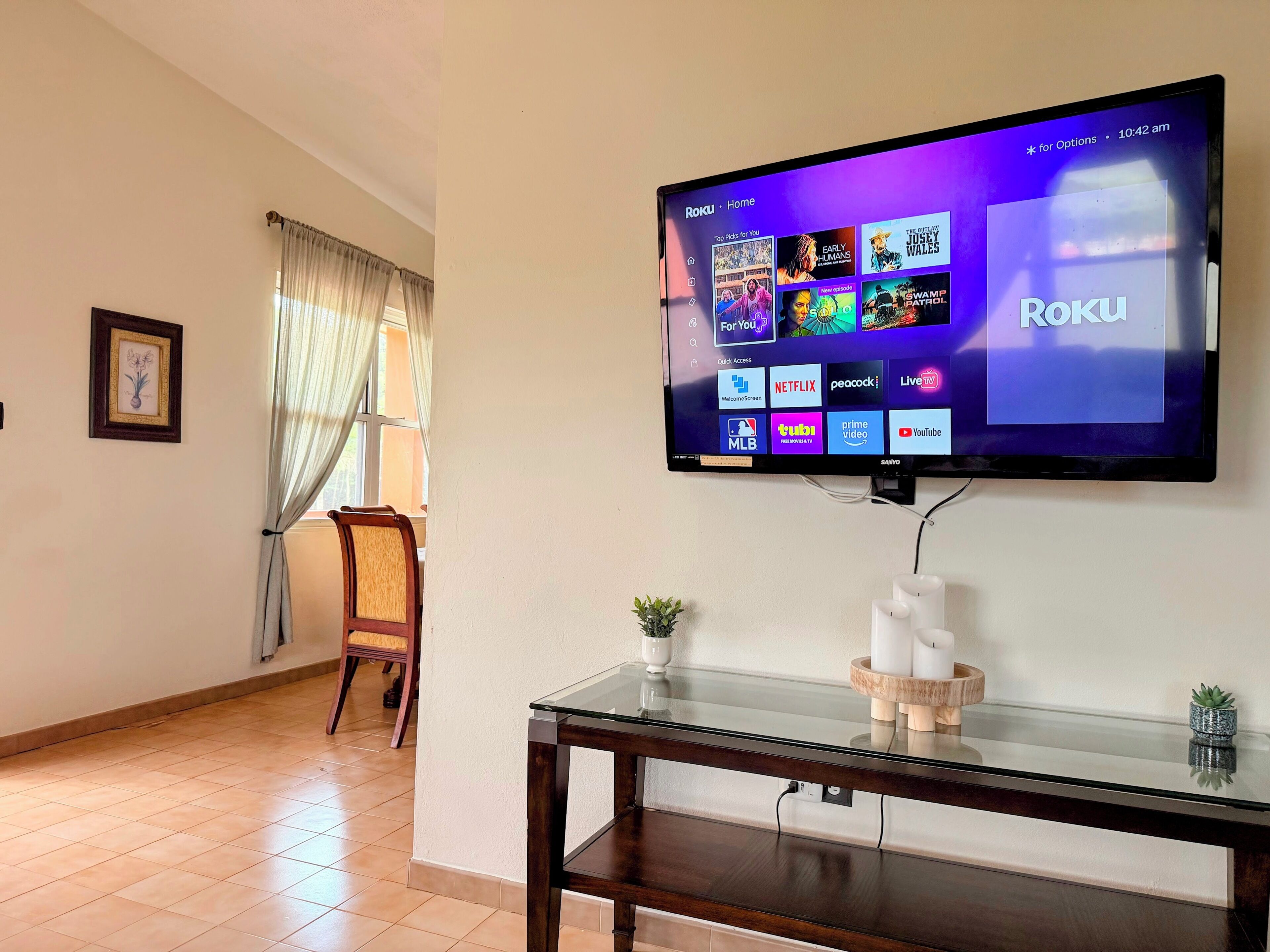A living room with a flat-screen TV mounted on the wall displaying the Roku interface.