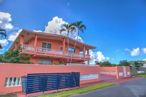 A pink two-story house with a balcony, a black fence, and a clear blue sky. - Countryside Villa with Pool, BBQ & El Yunque Views (Naguabo)