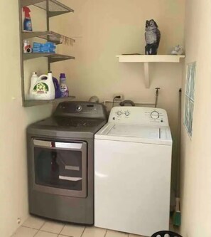 A laundry area with a washing machine and dryer, shelves with cleaning supplies, and a broom. - Countryside Villa with Pool, BBQ & El Yunque Views (Naguabo)