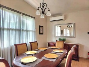 A dining room with a wooden table set for four, chairs, a chandelier, an air conditioning unit, and a mirror on the wall. - Countryside Villa with Pool, BBQ & El Yunque Views (Naguabo)