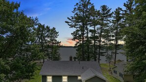 A two-story house with a dark green exterior, a small porch, and a view of a lake at sunset. - Cozy Private Lakefront Cottage, Pet-Friendly with Private Dock (Kalkaska)