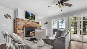 A living room with a fireplace, a television mounted above it, a ceiling fan, and a large window offering a view of the outdoors. - Cozy Private Lakefront Cottage, Pet-Friendly with Private Dock (Kalkaska)