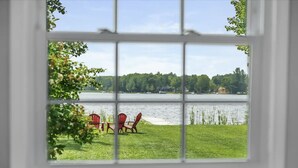 A window with a view of a lake, a grassy area, and red chairs. - Cozy Private Lakefront Cottage, Pet-Friendly with Private Dock (Kalkaska)