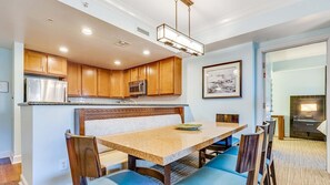 Een moderne keuken met houten kasten, een roestvrijstalen koelkast en een magnetron. Er staat een eettafel met blauwe stoelen en een bank.