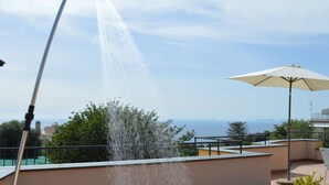Eine Dachterrasse mit Wasserspiel, weißem Sonnenschirm und Blick auf Meer und Berge.