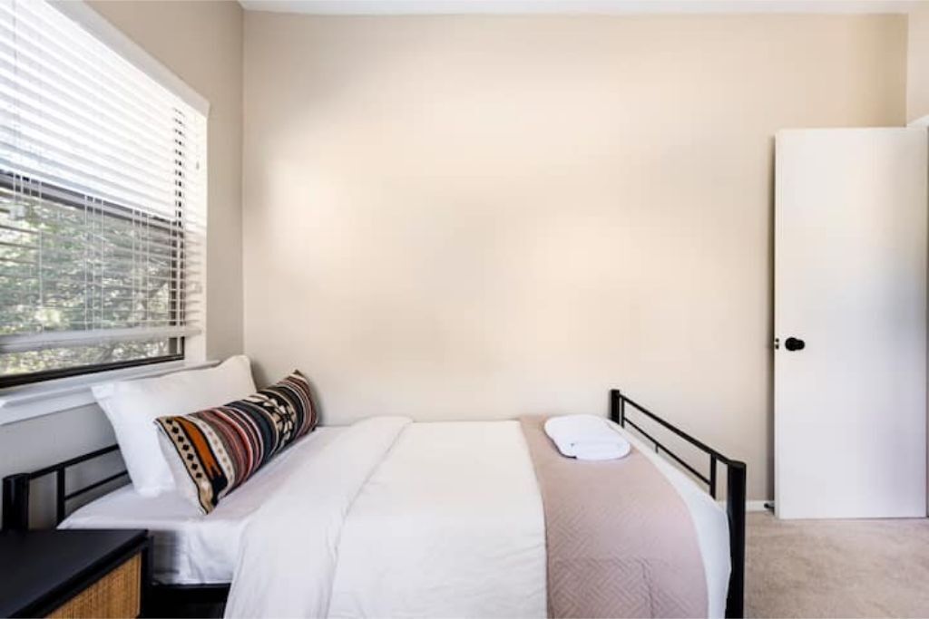 A bedroom with a bed, a window with blinds, a door, and a nightstand.