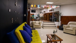 A lobby with a yellow sofa, a beige armchair, a wooden coffee table with a plant, and a wall with numbered clocks.