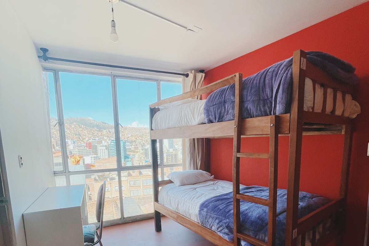 A bunk bed room with a desk and chair, large windows, and a view of buildings.