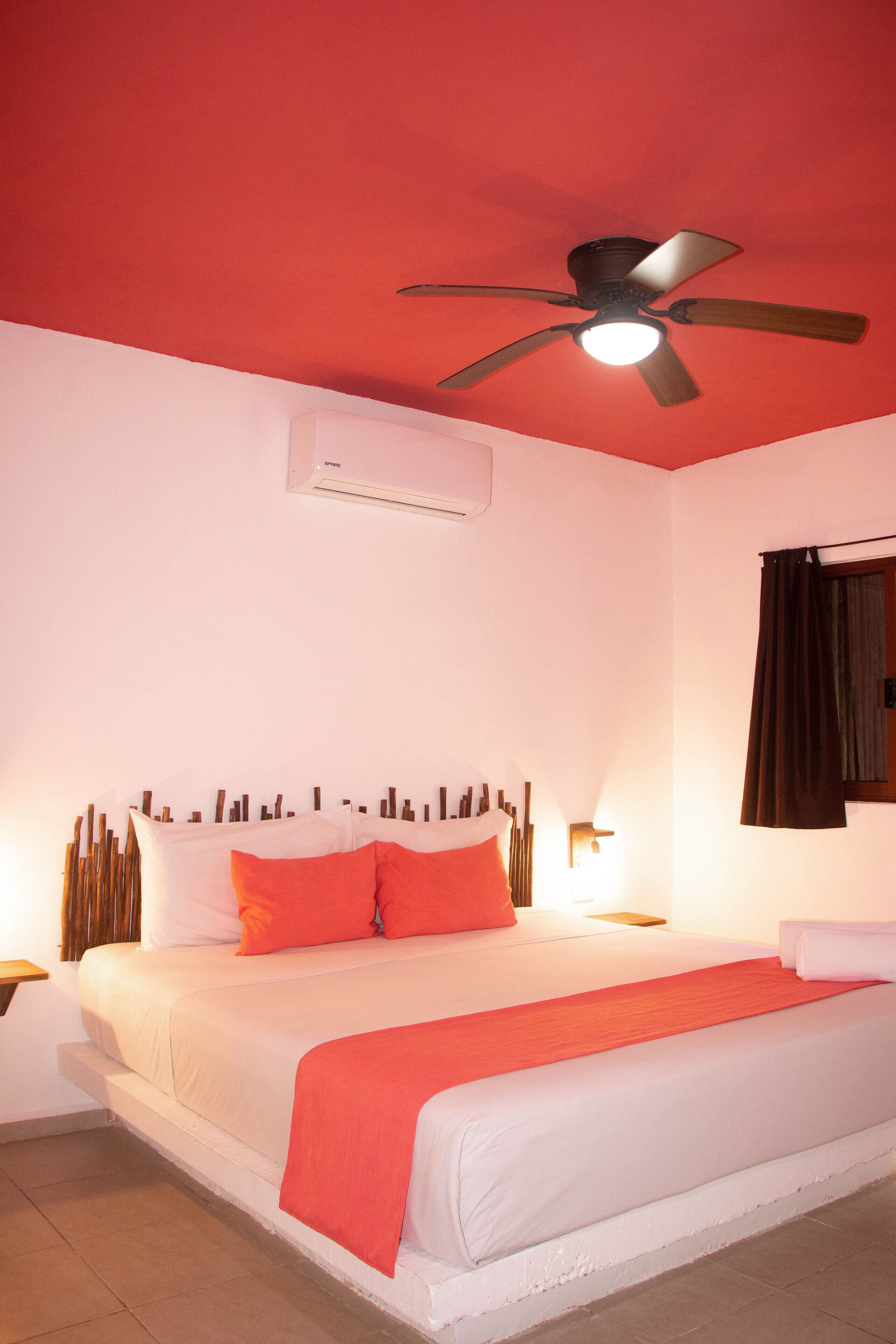 Una habitación de hotel con cama, ventilador de techo, aire acondicionado y ventana con cortinas.