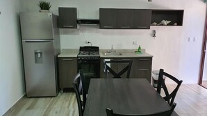 Une cuisine compacte équipée d’un réfrigérateur, d’une cuisinière et d’un évier. On y trouve une table à manger avec des chaises, ainsi qu’une plante en pot posée sur le plan de travail.