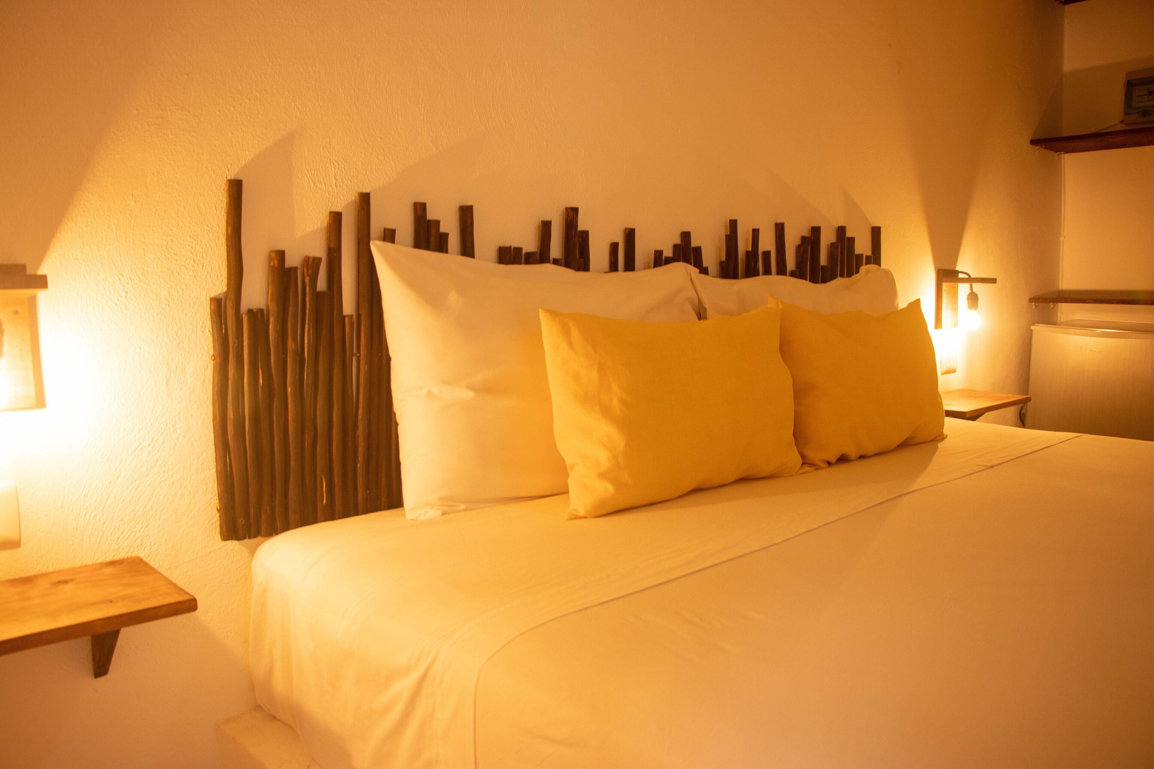 A hotel room with a bed, pillows, a wall-mounted light, and a wooden shelf.
