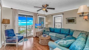 Ein Wohnzimmer mit Deckenventilator, einer Couch, einem Sessel, einem Fernseher und einem Balkon mit Blick.