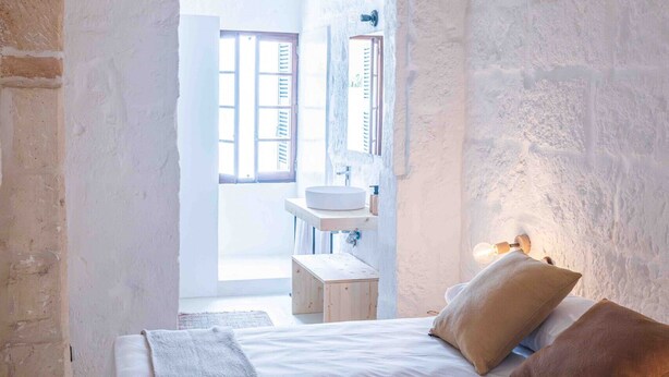 A bedroom with a wooden bed, a bench, a small bedside table, a wall-mounted air conditioner, and a window with shutters. - Pere Alcantara 40, 3 bedroom house, Ciutadella (Ciutadella de Menorca)