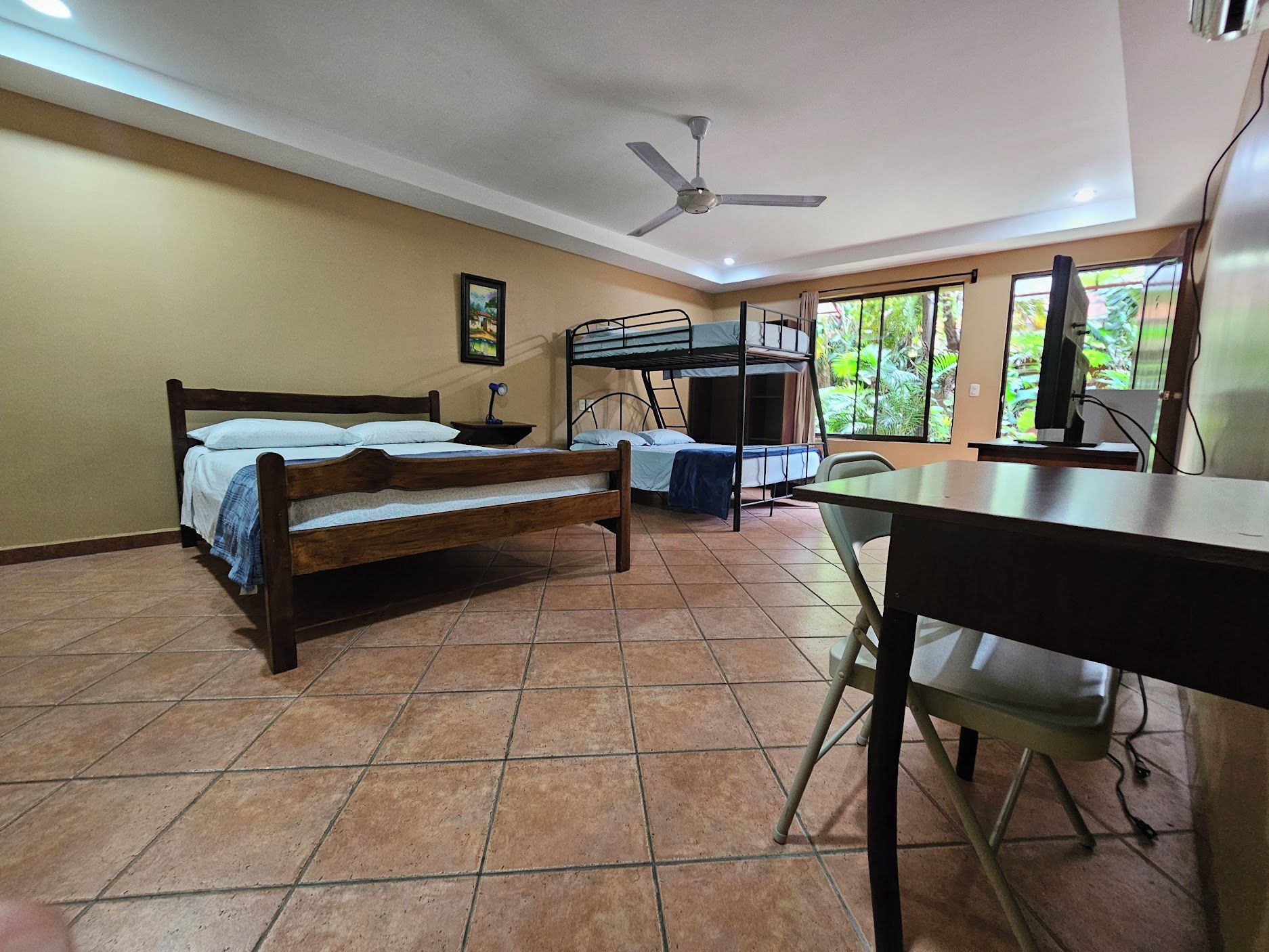 Una habitación con literas, un escritorio con computadora, una silla, un ventilador de techo y una ventana con vista a follaje.