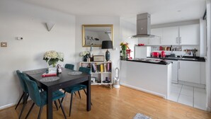 A modern kitchen with white cabinets, a black countertop, and a red kettle. There is a dining area with a dark table and teal chairs.