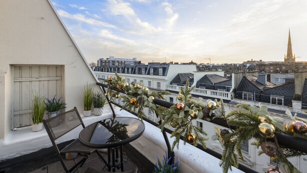 A balcony with a table and chairs, decorated with plants and Christmas ornaments, overlooking a cityscape with buildings and a church spire.