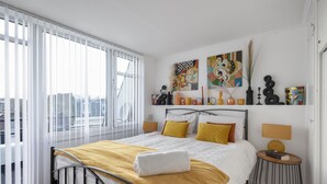 A modern bedroom with a large bed, white bedding, a yellow throw, and a city view through large windows.