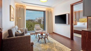 Una habitación de hotel moderna con un sofá, un sillón y una pequeña mesa con fruta. Un televisor está montado en la pared y hay una ventana grande con cortinas.