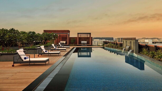 Piscina infinita en la azotea con sillas de descanso y una cascada, rodeada de edificios modernos y vegetación.