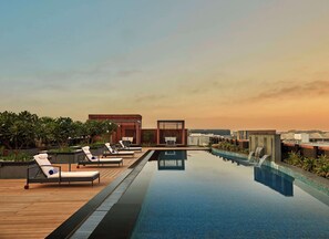 Piscina infinita en la azotea con sillas de descanso y una cascada, rodeada de edificios modernos y vegetación.