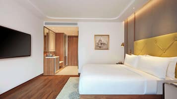 A modern hotel room with a large bed, a flat-screen TV mounted on the wall, wooden flooring, and a small bathroom with a sink and mirror.