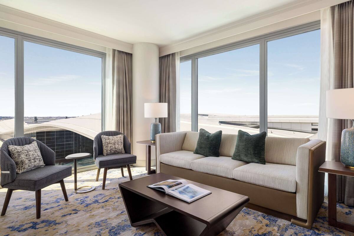A modern hotel room with a large window, a sofa, two armchairs, a coffee table, and a small side table with a lamp.