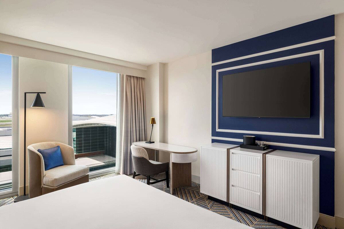 A hotel room with a large bed, a desk with a chair, a television mounted on the wall, and a view through the window.