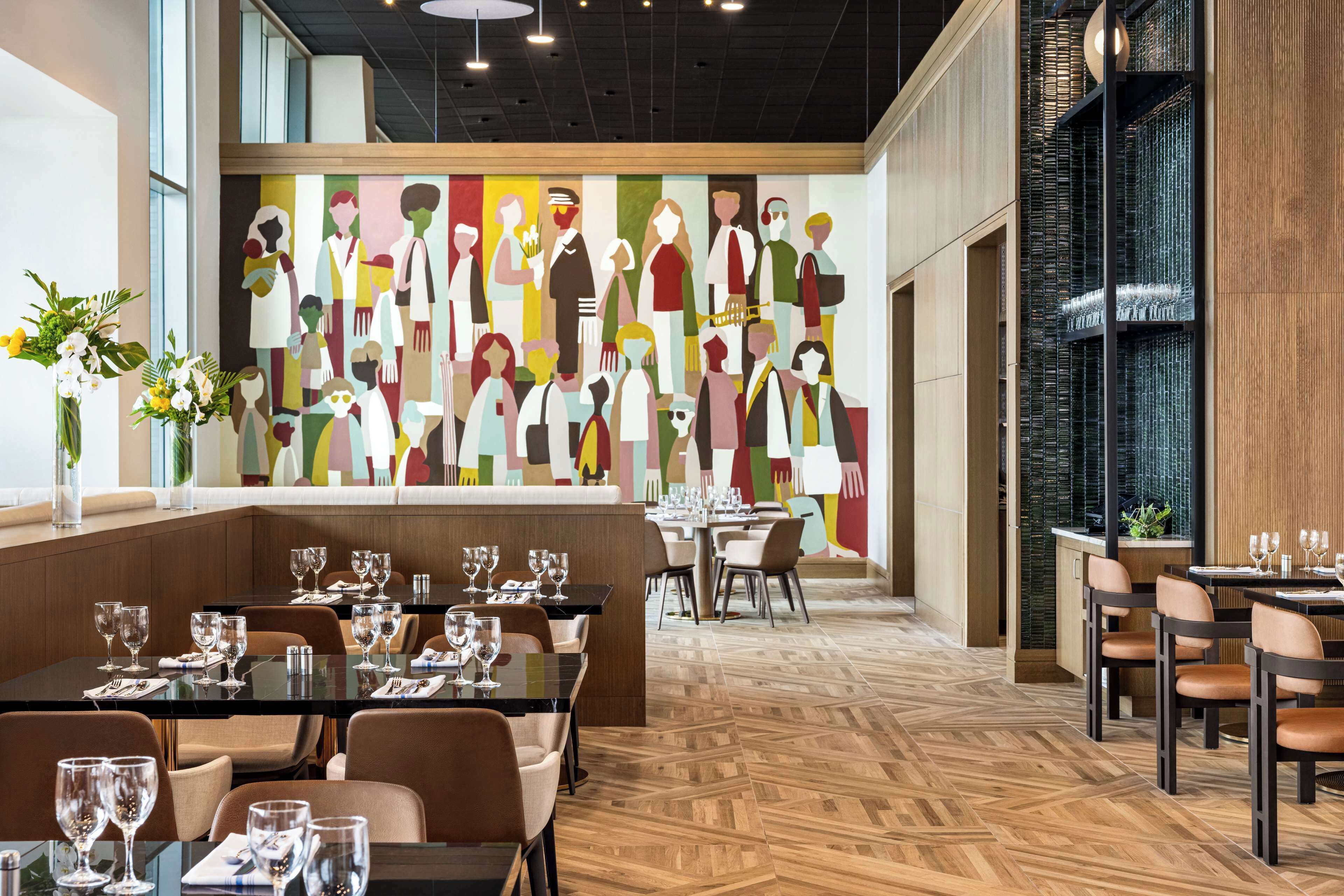 Een modern restaurant met een grote muurschildering van abstracte figuren, houten vloeren en netjes opgestelde tafels en stoelen.