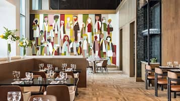 Un restaurant moderne, orné d’une grande fresque murale représentant des figures abstraites, doté d’un plancher en bois et de tables et de chaises disposées avec soin.