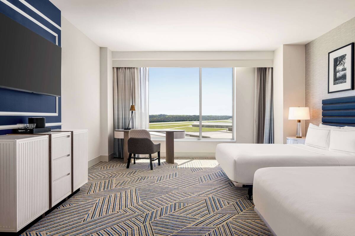A hotel room with two beds, a desk, a chair, a TV, and a large window with a view of a golf course.