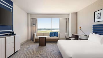 A hotel room with a large bed, a sofa, a small table, a TV, and a view of the airport.