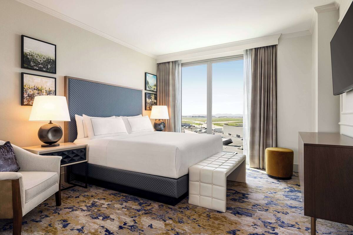 A hotel room with a large bed, a desk, a chair, a bench, and a TV.