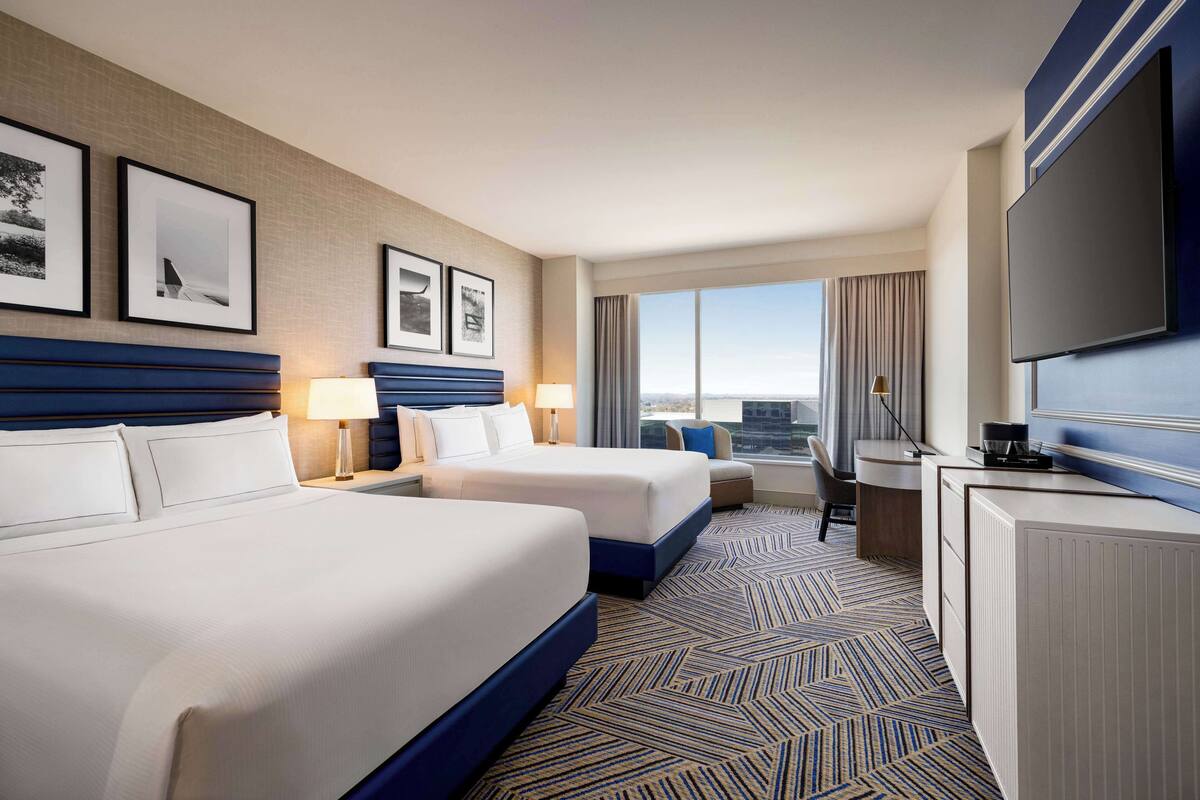 A hotel room with two beds, a desk, a TV, and a large window with a view.