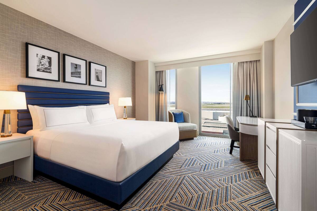 A hotel room with a large bed, a desk, a chair, and a TV. There are framed pictures on the wall and a view of the city from the window.
