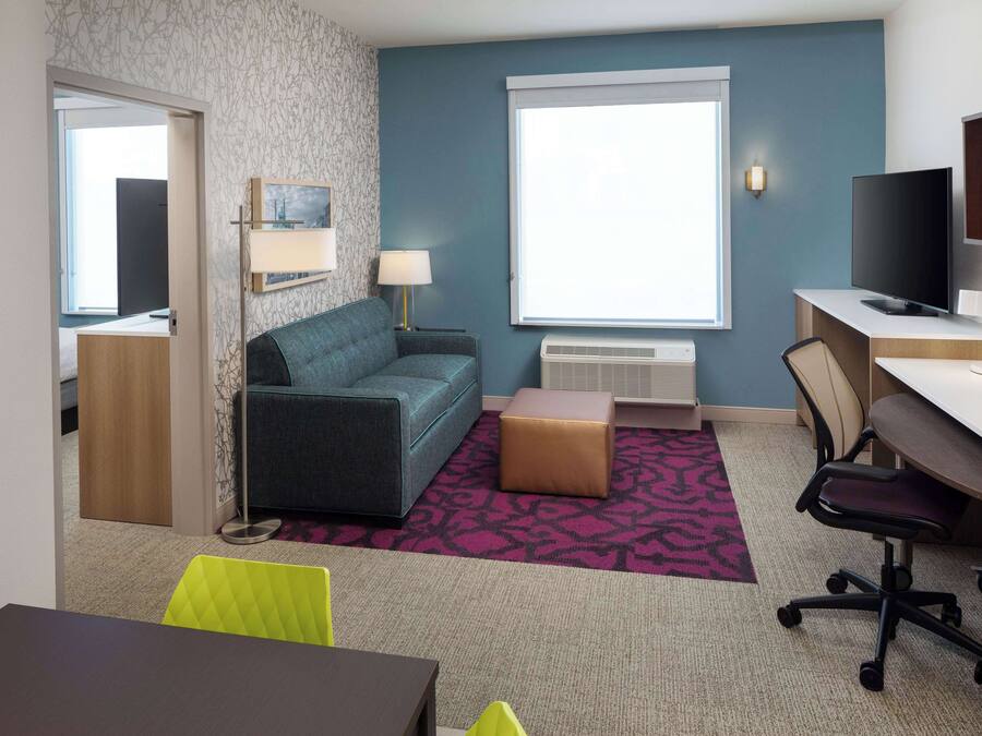A modern hotel room with a sofa, a desk, a television, and a window with blinds.