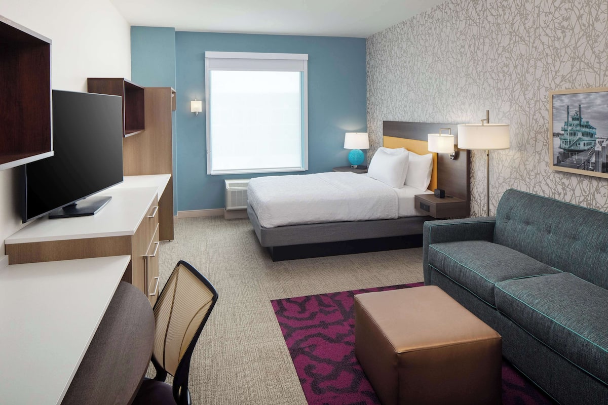 A hotel room with a bed, a desk with a flat-screen TV, a sofa, and a small table.