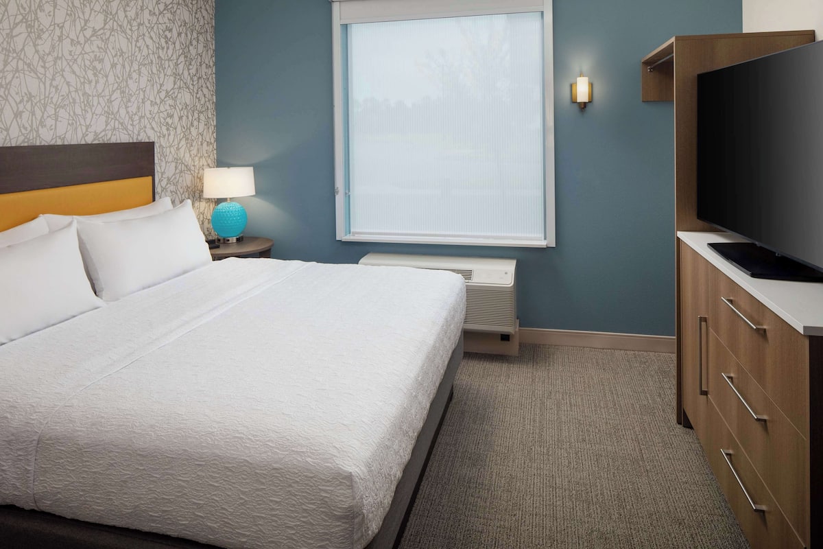A hotel room with a large bed, a TV, a nightstand with a lamp, and a window with blinds.