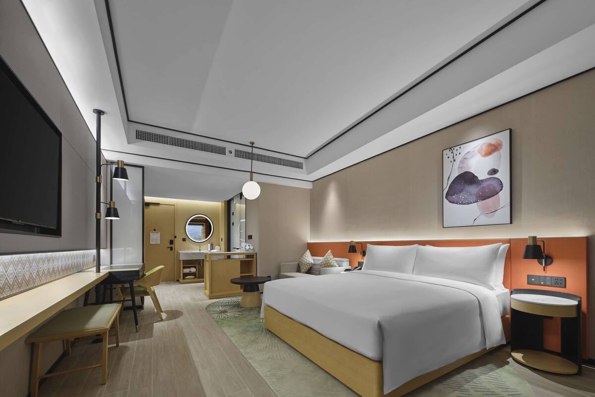 A modern hotel room with a large bed, a desk, a chair, a round mirror, and a painting on the wall.